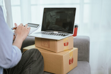 A young Asian female entrepreneur prepares packages and checks online orders from her home using a laptop, representing the concept of online shopping and a growing small SME startup