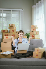 An Asian female entrepreneur prepares parcels and checks online orders from her home using her laptop and smartphone, reflecting the concept of online shopping and starting a SME business.