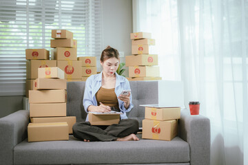 An Asian woman entrepreneur prepares parcels and manages her product orders via smartphone, ready to ship them to customers — reflecting the rise of online shopping and small business startups.