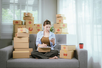 An Asian woman entrepreneur prepares parcels and manages her product orders via smartphone, ready to ship them to customers — reflecting the rise of online shopping and small business startups.