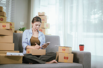 An Asian woman entrepreneur prepares parcels and manages her product orders via smartphone, ready to ship them to customers — reflecting the rise of online shopping and small business startups.