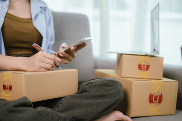 An Asian woman entrepreneur prepares parcels and manages her product orders via smartphone, ready to ship them to customers — reflecting the rise of online shopping and small business startups.