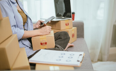 An Asian woman entrepreneur prepares parcels and manages her product orders via smartphone, ready to ship them to customers — reflecting the rise of online shopping and small business startups.