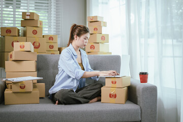A young Asian female entrepreneur prepares packages and checks online orders from her home using a laptop, representing the concept of online shopping and a growing small SME startup