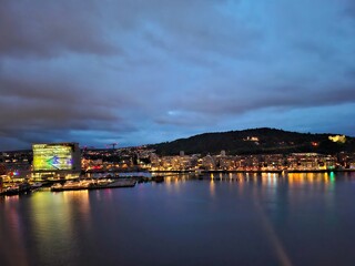 Norway oslo june 12 2024 Colorful city lights illuminate the waterfront under a cloudy evening sky, reflecting on calm water.