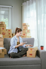A young Asian female entrepreneur prepares packages and checks online orders from her home using a laptop, representing the concept of online shopping and a growing small SME startup