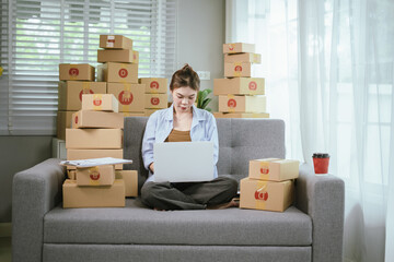 A young Asian female entrepreneur prepares packages and checks online orders from her home using a laptop, representing the concept of online shopping and a growing small SME startup