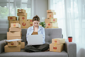 A young Asian female entrepreneur prepares packages and checks online orders from her home using a laptop, representing the concept of online shopping and a growing small SME startup