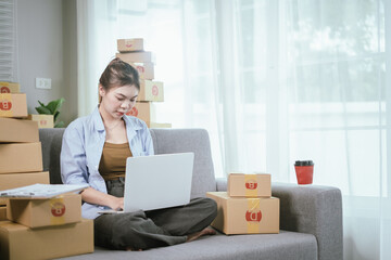 A young Asian female entrepreneur prepares packages and checks online orders from her home using a laptop, representing the concept of online shopping and a growing small SME startup