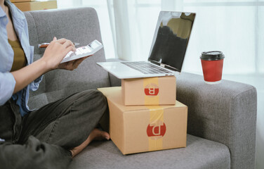 A young Asian female entrepreneur prepares packages and checks online orders from her home using a laptop, representing the concept of online shopping and a growing small SME startup
