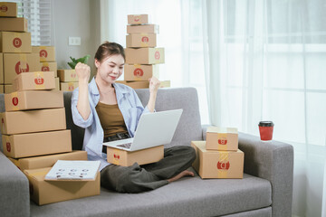 An Asian female entrepreneur is preparing parcels and checking online orders of her products to ship to customers using a laptop at home. A concept of online shopping and a small startup SME business.