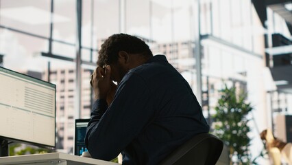 Worker feeling painful headache while working, suffering burnout while rushing to finish tasks before deadline. African american man in office having migraine after working overtime, camera A