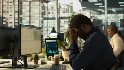Worker feeling painful headache while working, suffering burnout while rushing to finish tasks before deadline. African american man in office having migraine after working overtime, camera B