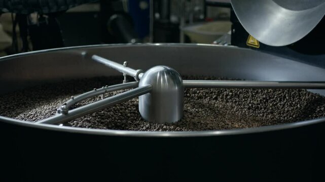 Professional coffee roasting machine rotating metal tool thoroughly mixing dark roasted coffee beans during industrial manufacturing process, showcasing detailed texture and preparation