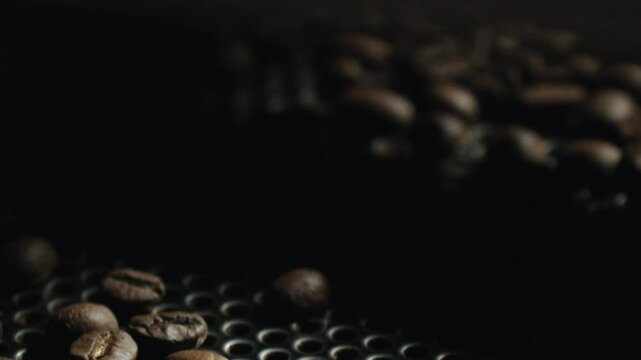 Roasted coffee beans gradually cooling and rolling across metal grid surface, showcasing professional roasting equipment and artisanal production process within industrial coffee workshop