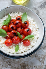 Caprese with cottage cheese, roasted cherry tomatoes and fresh basil in a grey bowl, vertical shot on a brown granite surface, middle closeup