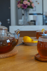 Glass teapot and cups with hot tea and lemon on wooden table in bright living room. Warm light and cozy interior. Concept of wellness and relaxation