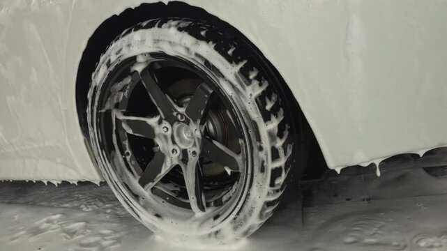 A smooth panning close-up of a tire blanketed in thick soap foam, capturing the vibrant texture and satisfying cleaning process during a car wash.