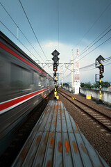 A blur of a train passes a station platform with tracks and railway signals under a cloudy sky,...