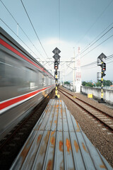 A blur of a train passes a station platform with tracks and railway signals under a cloudy sky,...