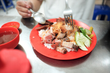 A diner enjoys a plate of Nasi Campur, a flavorful Peranakan Chinese-Indonesian dish featuring assorted roasted pork and egg, served in a bustling eatery