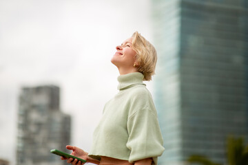 Mature woman with blond hair looking up and smiling while holding phone outdoors