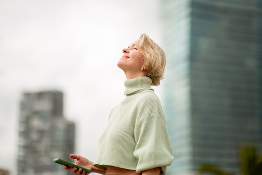 Mature woman with blond hair looking up and smiling while holding phone outdoors