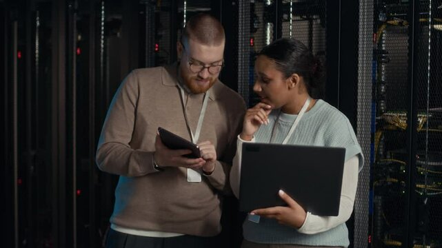 Medium shot of two intercultural IT specialists having discussion using digital devices in server room - Powered by Adobe