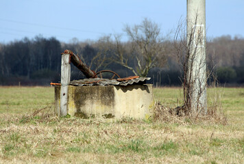 Weathered concrete well with a rusted metal pipe and corrugated cover stands in a grassy open...
