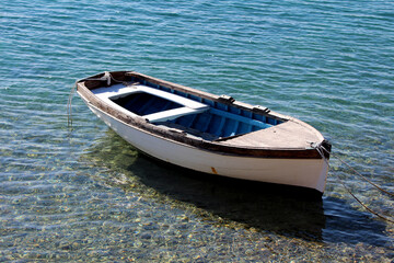 White and blue wooden rowboat with dark trim floats in shallow, crystal-clear water, tethered by ropes at the bow and stern, revealing pebbles beneath and gentle ripples in bright sunlight