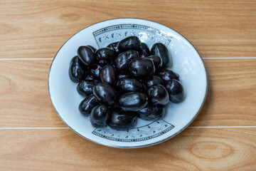 Fresh black plums on white plate, wooden background. Delicious Java plum, Jamun fruit (Syzygium...