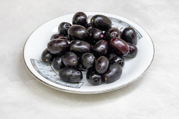 Ripe fresh black Java plums in white plate. Healthy jamun fruit, also known as Malabar plum, jambul, jambolan.