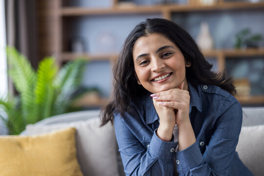 Portrait of a positive young smiling Indian woman sitting on the sofa at home, folding her hands together and looking confidently at the camera
