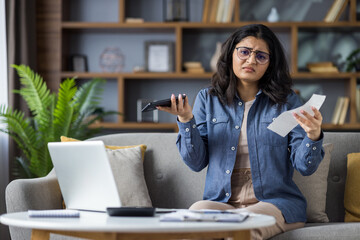 Portrait of upset young Indian woman sitting on sofa at home, holding documents, bills and mobile phone, spreading her arms at camera in disappointment