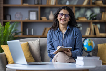 Portrait of a smiling young Indian female teacher sitting at home on the couch in front of the...