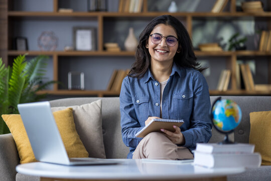 Portrait of a smiling young Indian female teacher sitting at home on the couch in front of the screen, holding notebooks in her hands and talking on the video