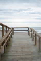 Obraz premium Wooden bridge on the beach under a cloudy sky. Coastal landscape with sand, natural textures, and a moody atmosphere, evoking solitude and connection with nature.