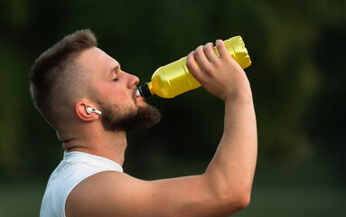 A young fit man with closed eyes listening to music on wireless headphones and drinking water or...