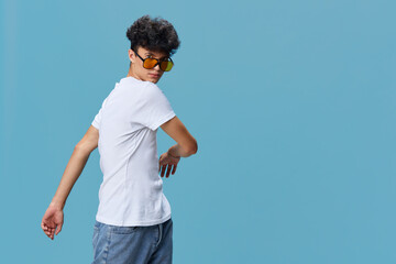 Young man wearing white t-shirt and yellow sunglasses poses confidently against blue background, showcasing casual style and relaxed attitude with natural lighting.