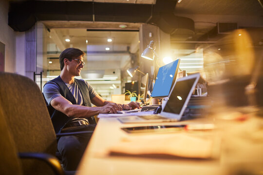 Young professional man working late at modern office desk with multiple screens and warm lighting