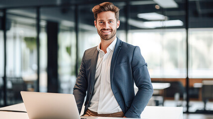 A confident young man in a suit smiles while sitting at a desk with a laptop in a modern office setting.
