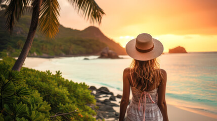 A woman in a sunhat stands on a tropical beach at sunset, overlooking the calm ocean and lush green hills.