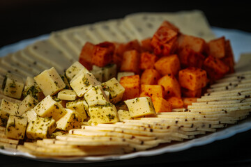 Close-up of a cheese platter featuring spiced and herbed cheese cubes, and thinly sliced cheese arranged on a white plate over a dark surface.