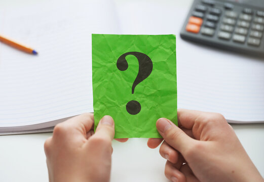 A student  holding a green paper note with a question mark on it in his hands over an open notebook and a calculator. Close up.