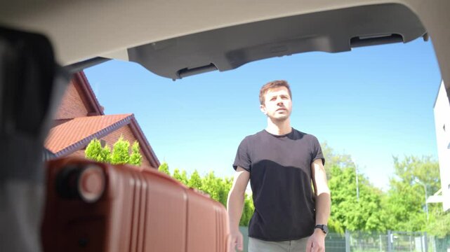 Man placing orange rolling suitcase into open vehicle trunk, preparing for journey or vacation on sunny day. Inside view of traveler loading luggage in car. Travel or weekend road trip concept
