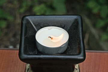 one burning white round small paraffin candle stands in a ceramic brown black jug on a table on a...