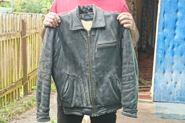 hands of man in red clothes holding old black leather jacket with fur on summer street