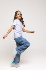 Cheerful genius positive little girl with curly hair in white t-shirt, blue jeans and sneakers has fun and making a step on white background. Happy childhood concept. Copy space. High quality photo