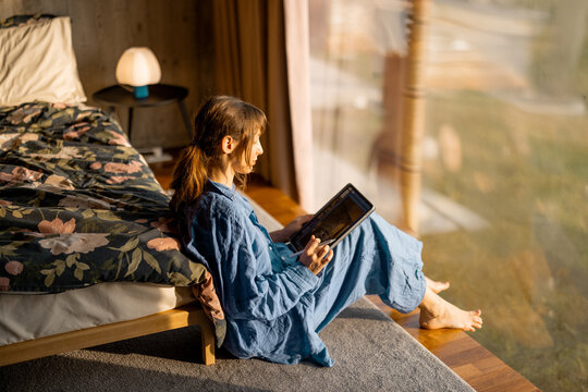 A woman in blue linen loungewear sits by a panoramic window, reading on a tablet. Soft morning light, cozy atmosphere, and a peaceful connection with nature