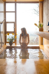 A woman enjoys a serene bath in a wooden tub by a panoramic window. Surrounded by flowers and soft...
